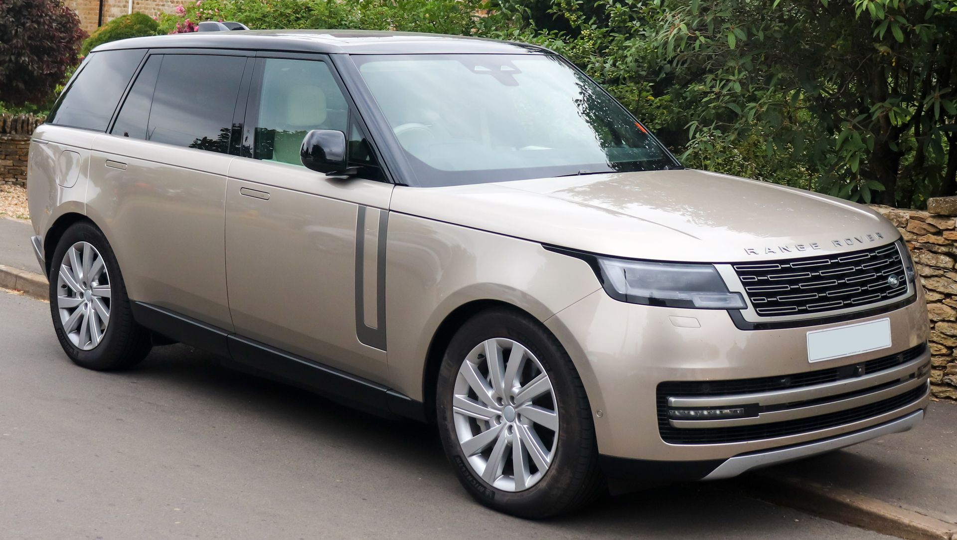 Gold Range Rover SUV parked on a street with a stone wall and greenery.