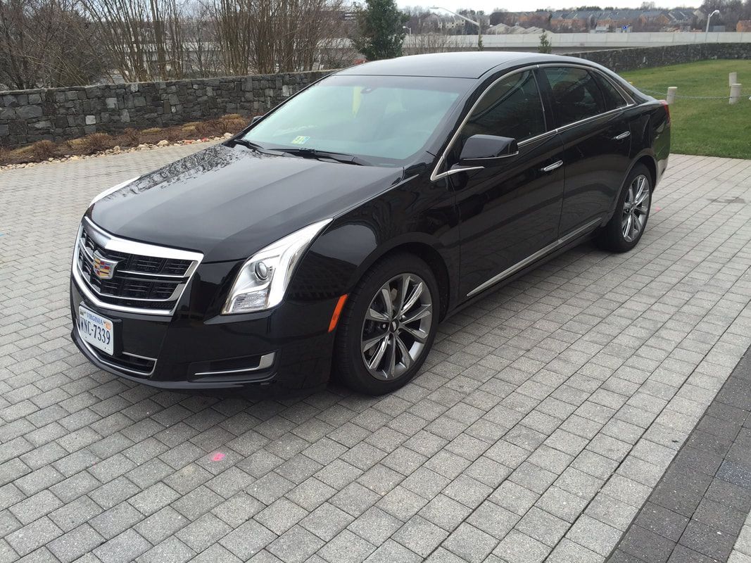 Black Cadillac sedan parked on a brick driveway, in front of a stone wall.