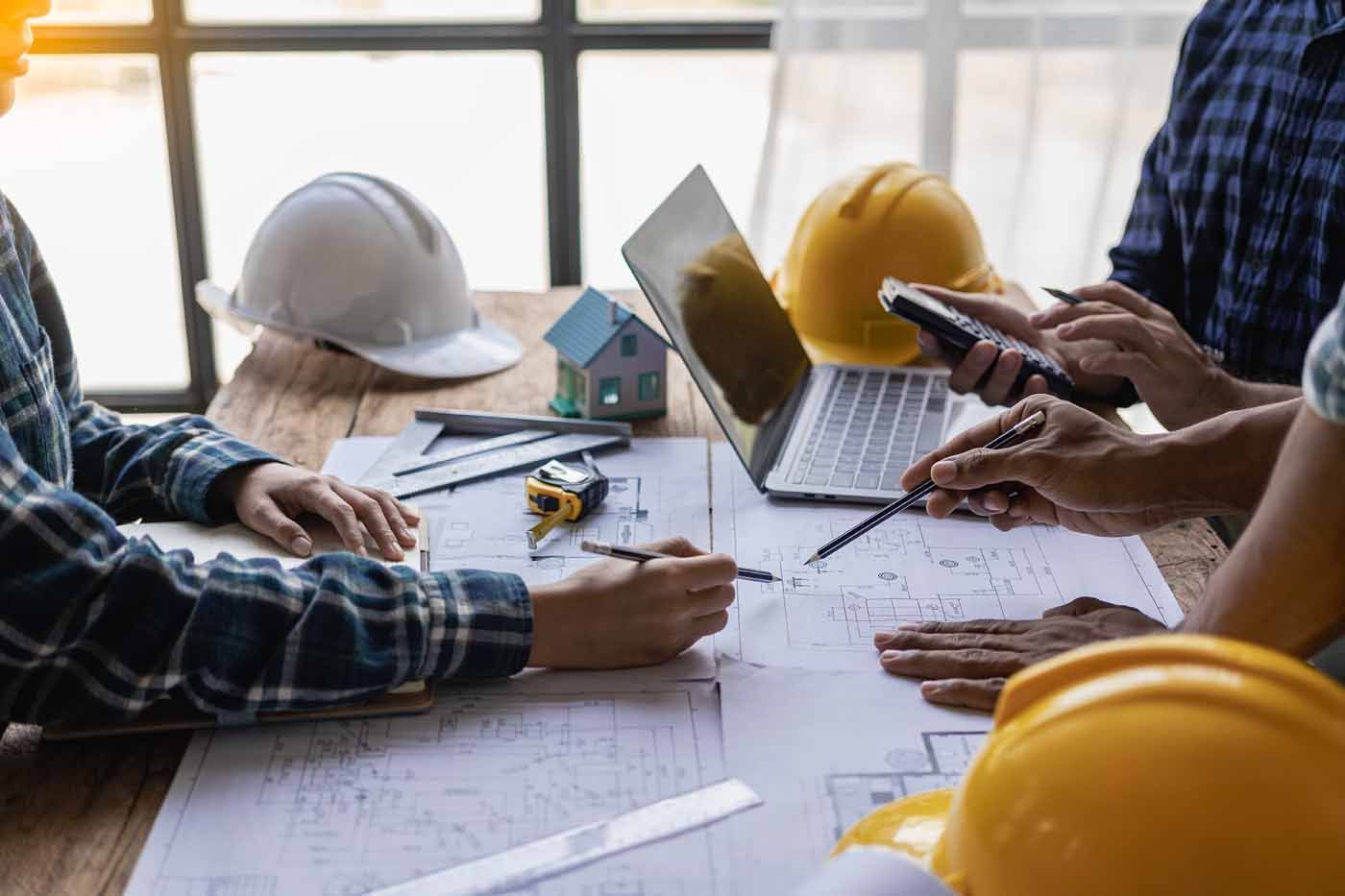A group of construction workers are sitting around a table with hard hats and a laptop.