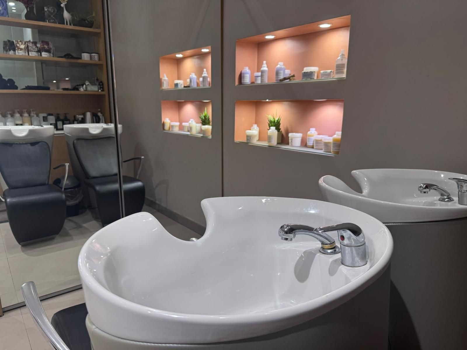 Salone moderno con lavatesta bianchi, poltrone grigie e mensole a parete illuminate che espongono i prodotti.