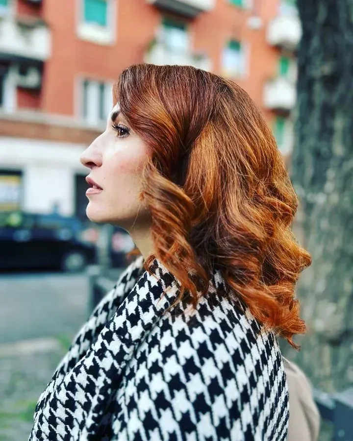 Profilo laterale di una donna con capelli ramati ondulati che indossa un cappotto a quadri pied de poule bianco e nero all'aperto.