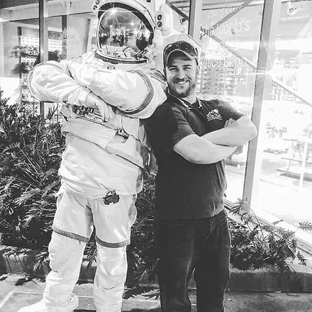 Man with crossed arms poses next to an astronaut in a full spacesuit near a window.