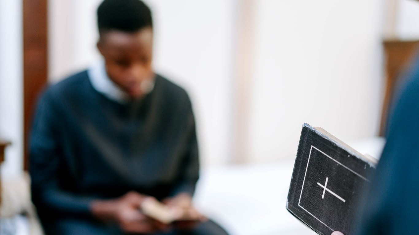 Several hands are held together in prayer over a Bible resting on a wooden table.