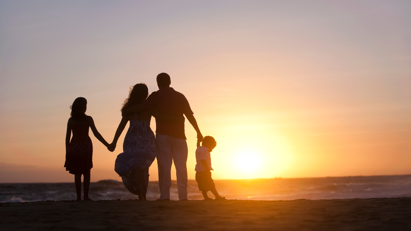 Silhouetted family holding hands on a beach at sunset, with warm orange sky and ocean horizon.