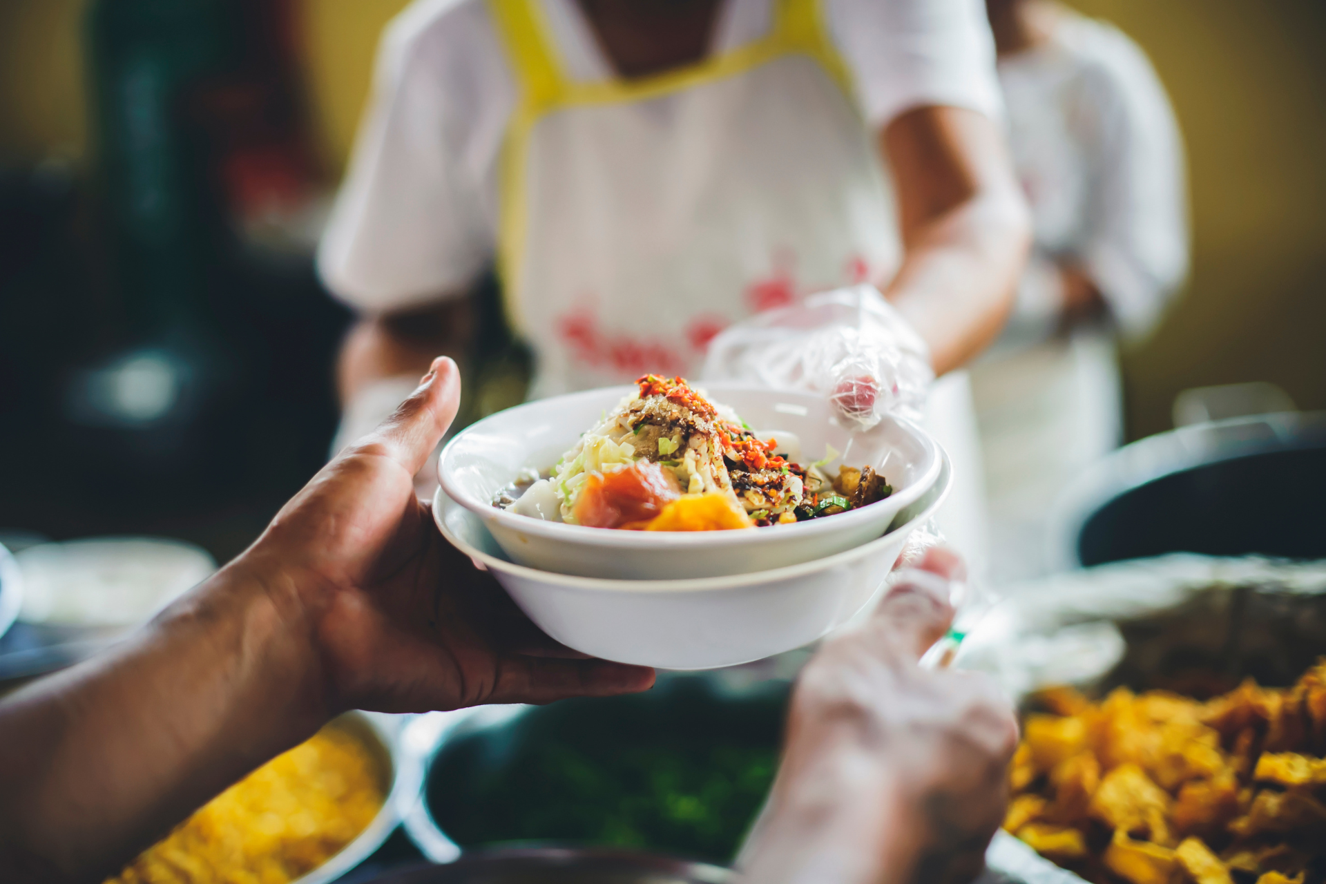 Hand holding a bowl of rice and vegetables at a food stall, with blurred prepared dishes in the background