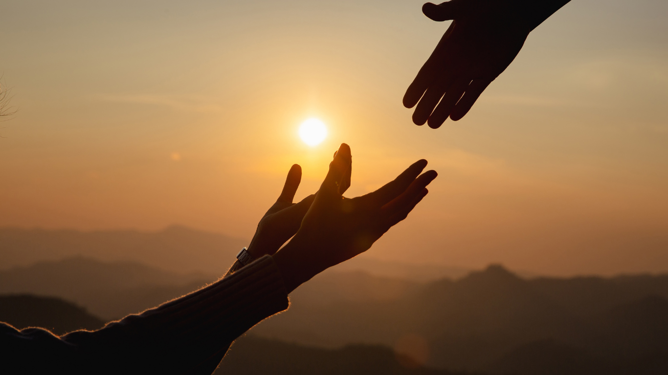 Two hands reaching toward each other against a sunset over distant mountains