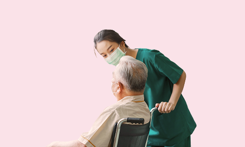 Caregiver in green scrubs leaning toward seated patient in wheelchair against a pink background