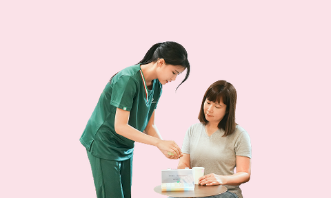 Healthcare worker in green scrubs examining a document at a table with patient against a pink background