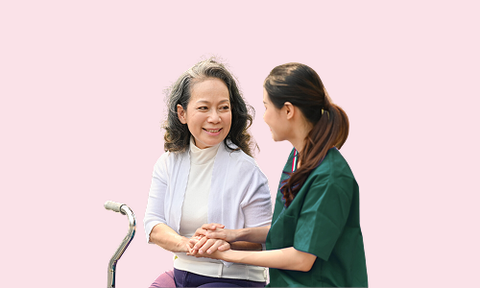 Caregiver in medical scrubs smiling and talking with elderly holding a cane against a pink background