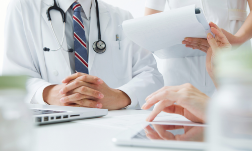 Doctor and patient discussing paperwork at a desk, with a stethoscope and laptop visible.