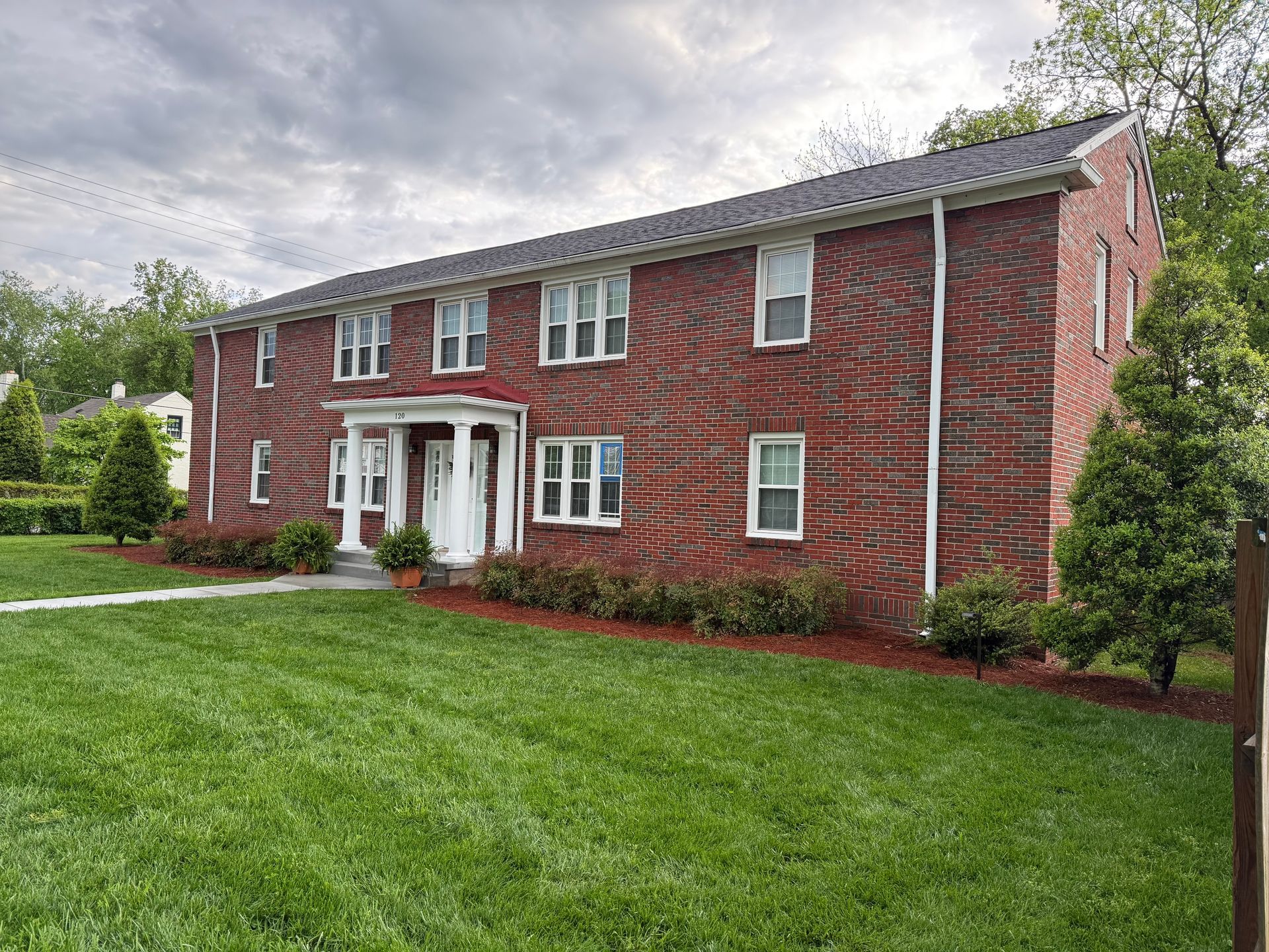 White two-story house with green shutters and a front stoop; a black lamp post stands in front. Two-story red brick apartment building with white columns and trim, green lawn, cloudy sky.
