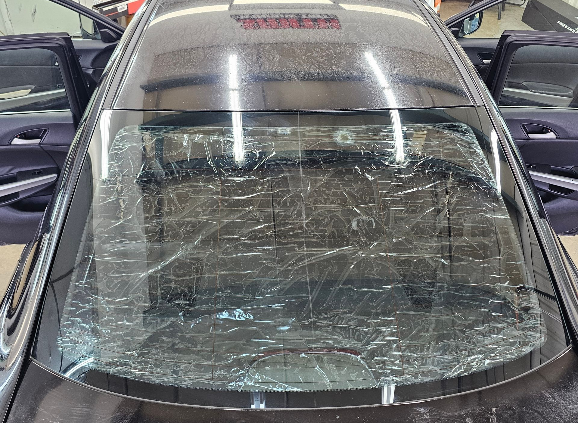 Black car's rear window covered in scratched, clear film, with open doors in a shop.