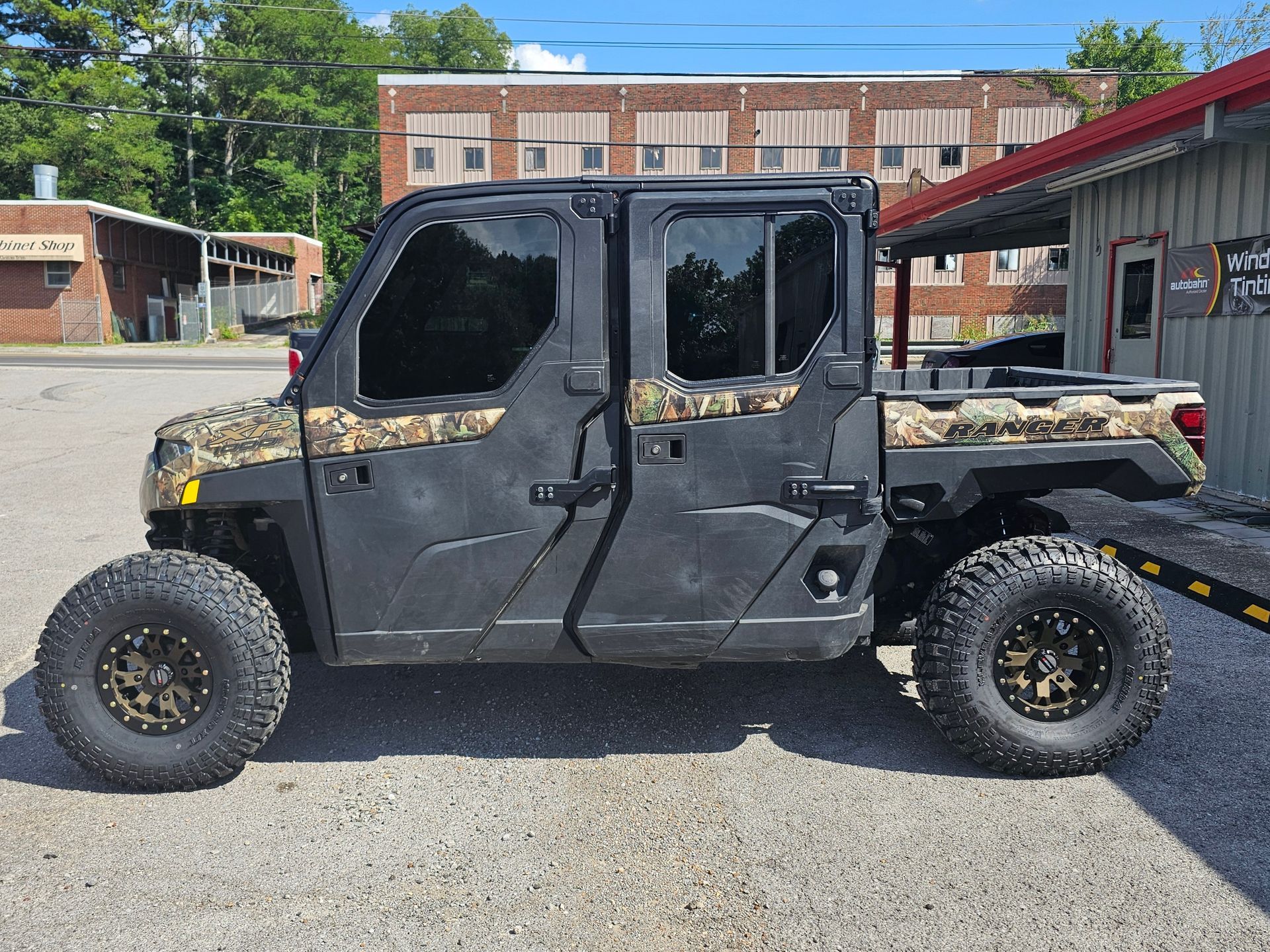 Black and camo side-by-side utility vehicle with large tires parked on pavement, in front of buildings.