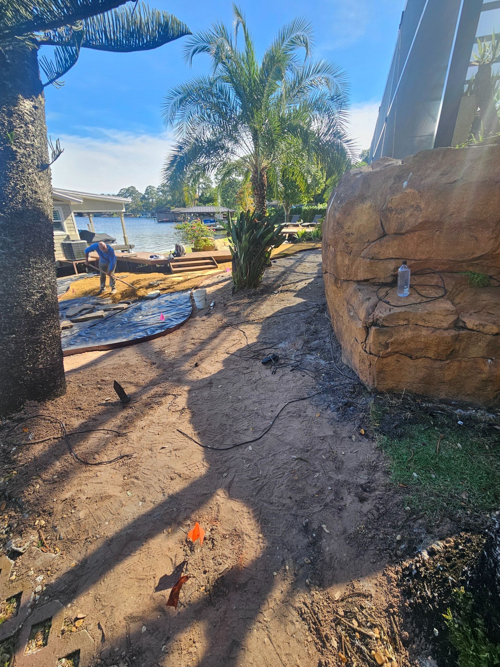 A dirt path winds past a rock wall and towards a person near a body of water under a sunny sky.