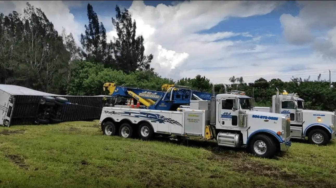 Two white tow trucks parked on grass beside a black trailer under a cloudy sky