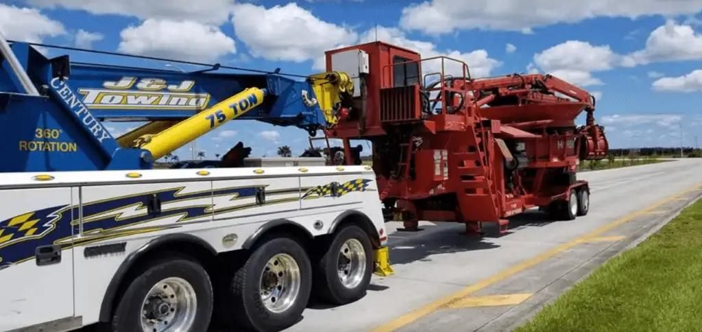 Large red industrial machine on a trailer beside a blue-and-white truck on a road under a cloudy sky