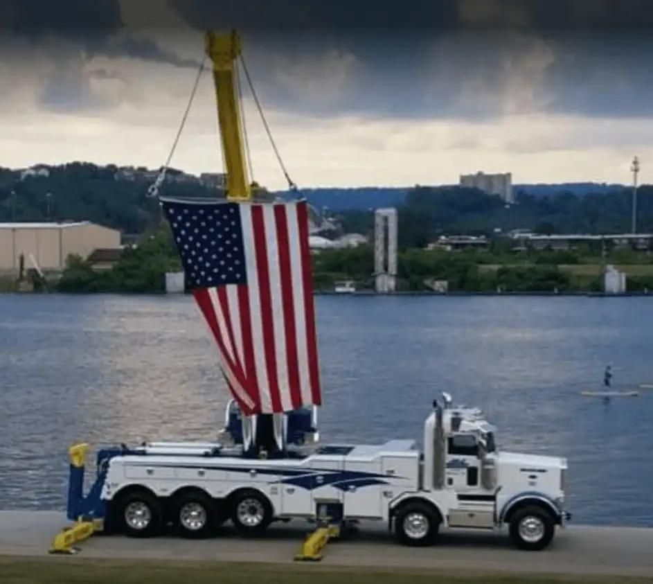 Large American flag hanging from a white tow truck by a waterfront under a cloudy sky