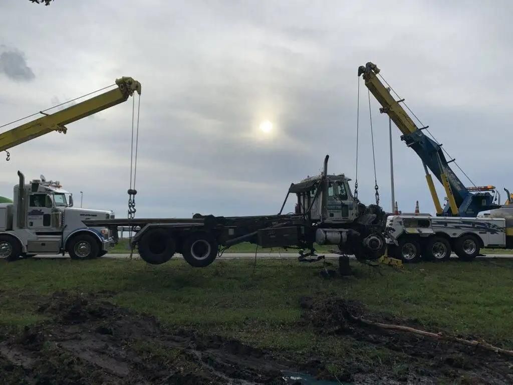 Two tow trucks lifting a long flatbed trailer in a muddy field under a cloudy sky