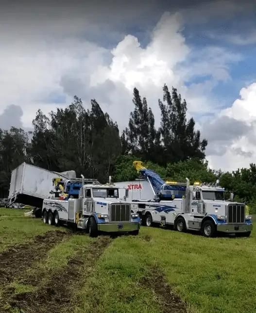 Two white tow trucks and a dump truck on a grassy field under cloudy skies