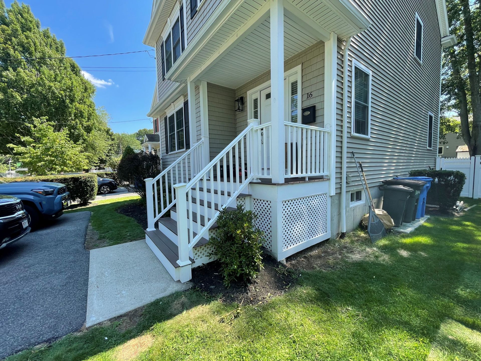 Front porch of a gray house with white railings and steps, beside a sidewalk and grassy yard.