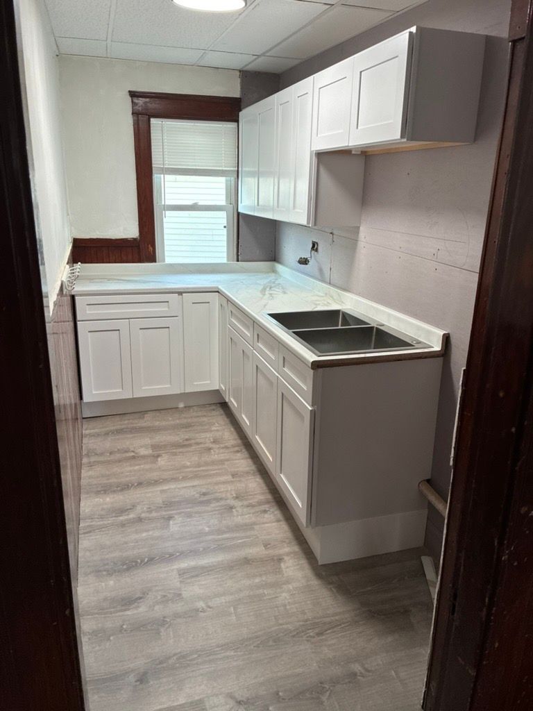 Empty white kitchen with cabinets, beige countertop, and a window letting in sunlight