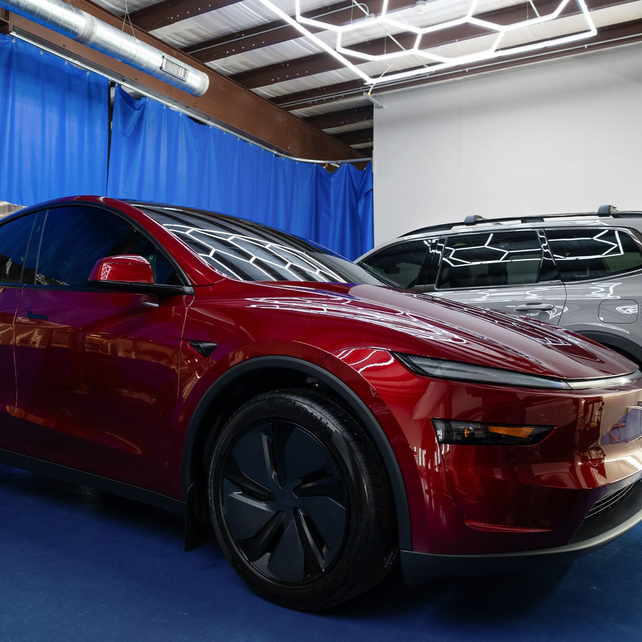 Red electric car parked in a garage next to a gray car. Blue curtains and bright lights are visible.