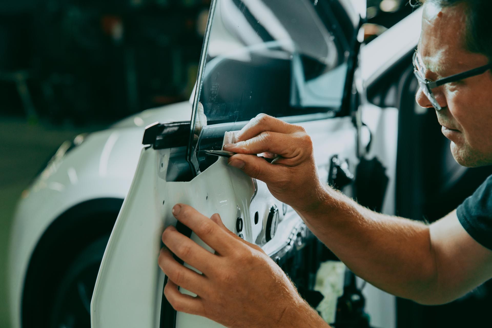 Man repairing a car door in a garage, using a small tool. White car, close-up.