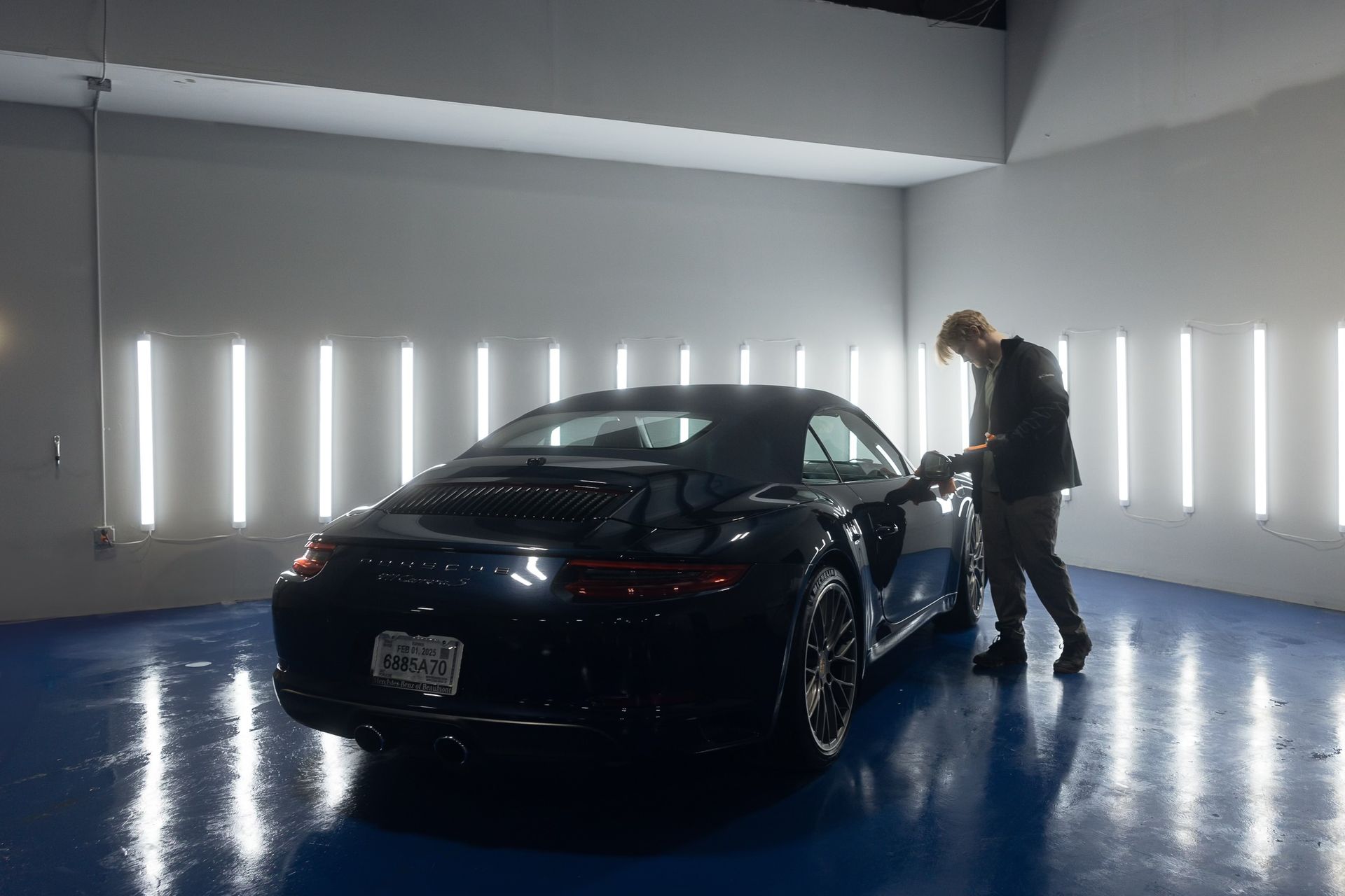 Man polishing a black Porsche in a well-lit garage with vertical light panels.