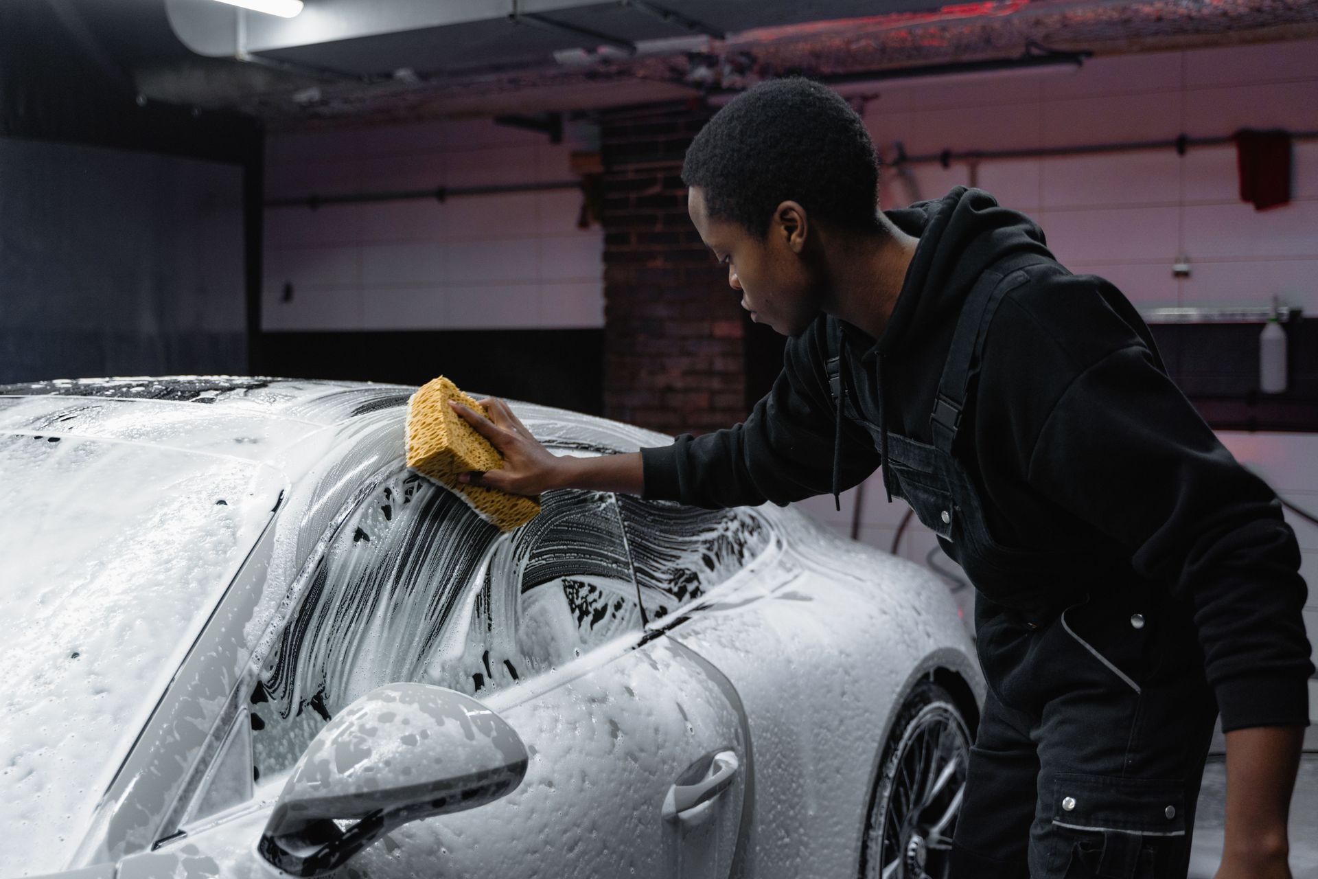 Person washing a silver sports car with a yellow sponge in a garage