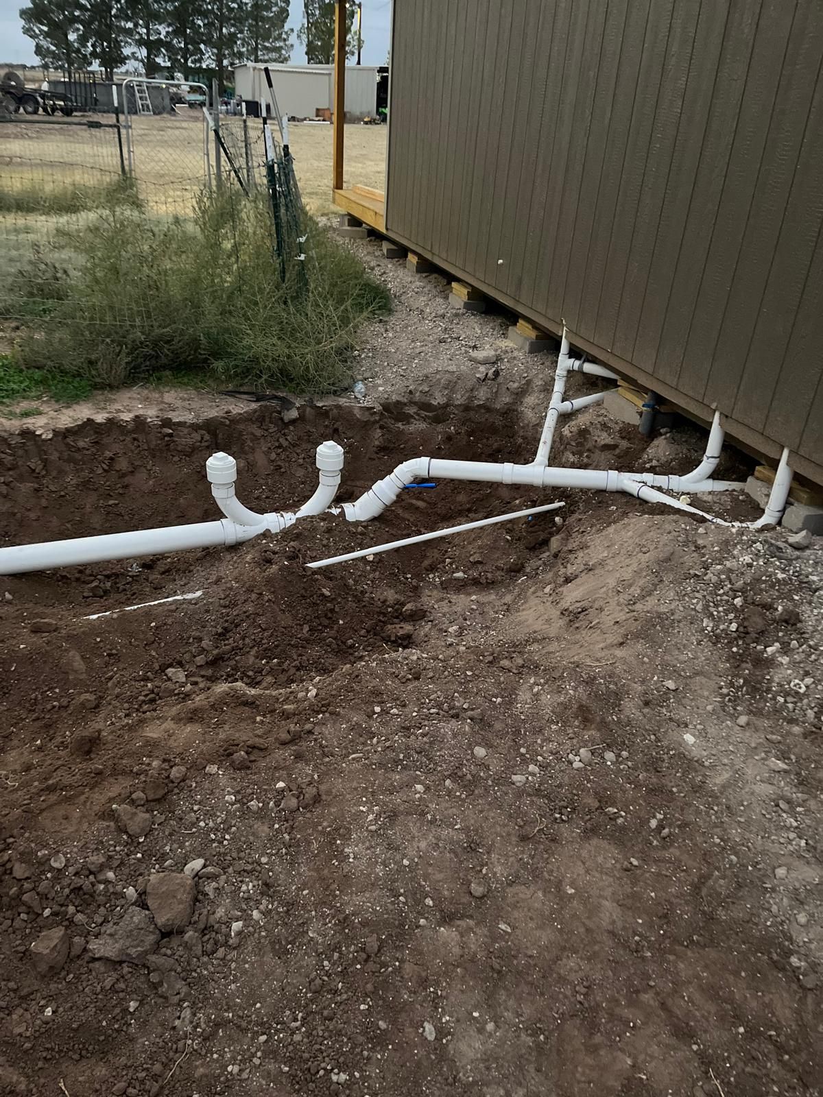 Pipes laid in a trench next to a tan building and dirt, likely for plumbing or drainage.
