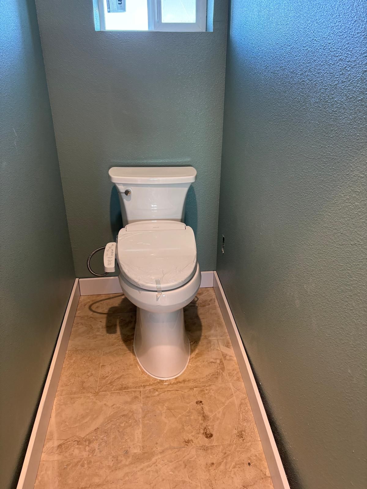 Toilet in a narrow bathroom alcove with a small window above. The walls are green and the floor has tile.