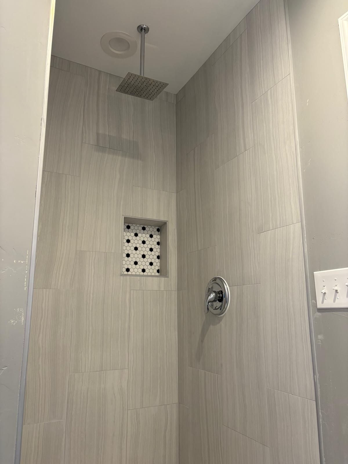Shower stall with light gray tile walls and a square shower head. Includes a built-in shelf.