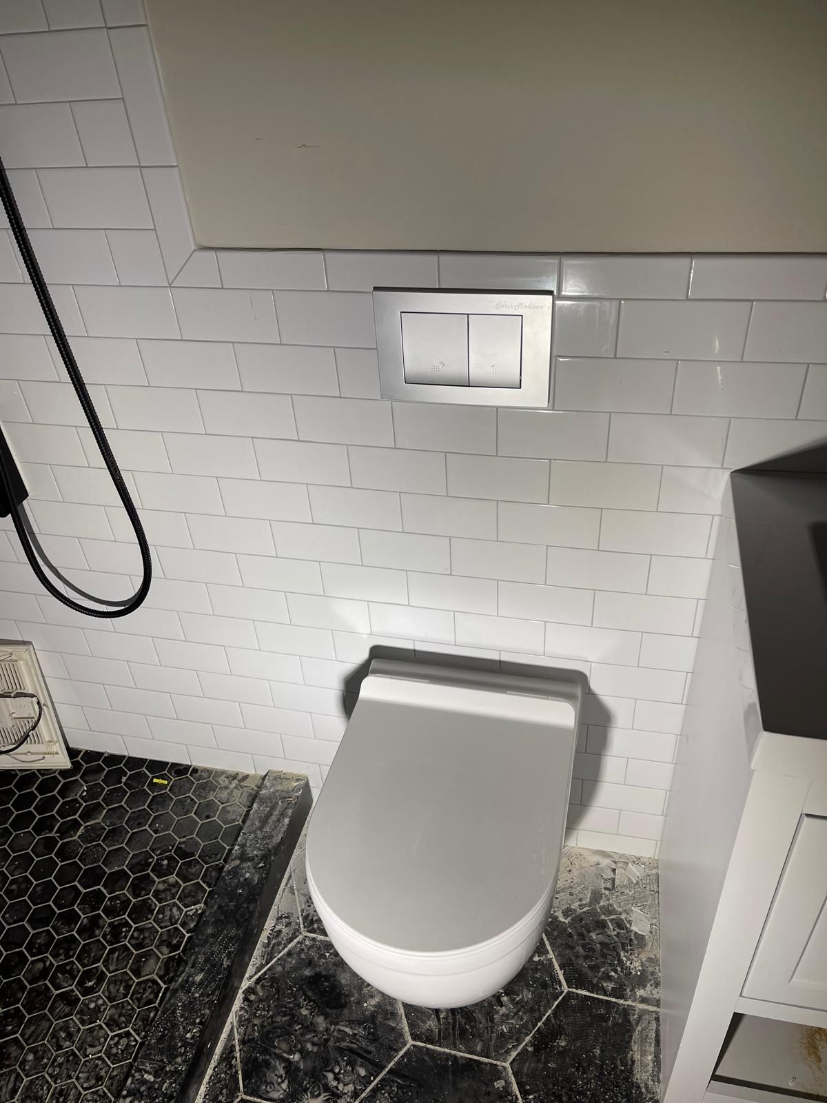 Toilet with wall-mounted flush buttons on white tiled wall; black and white patterned floor.
