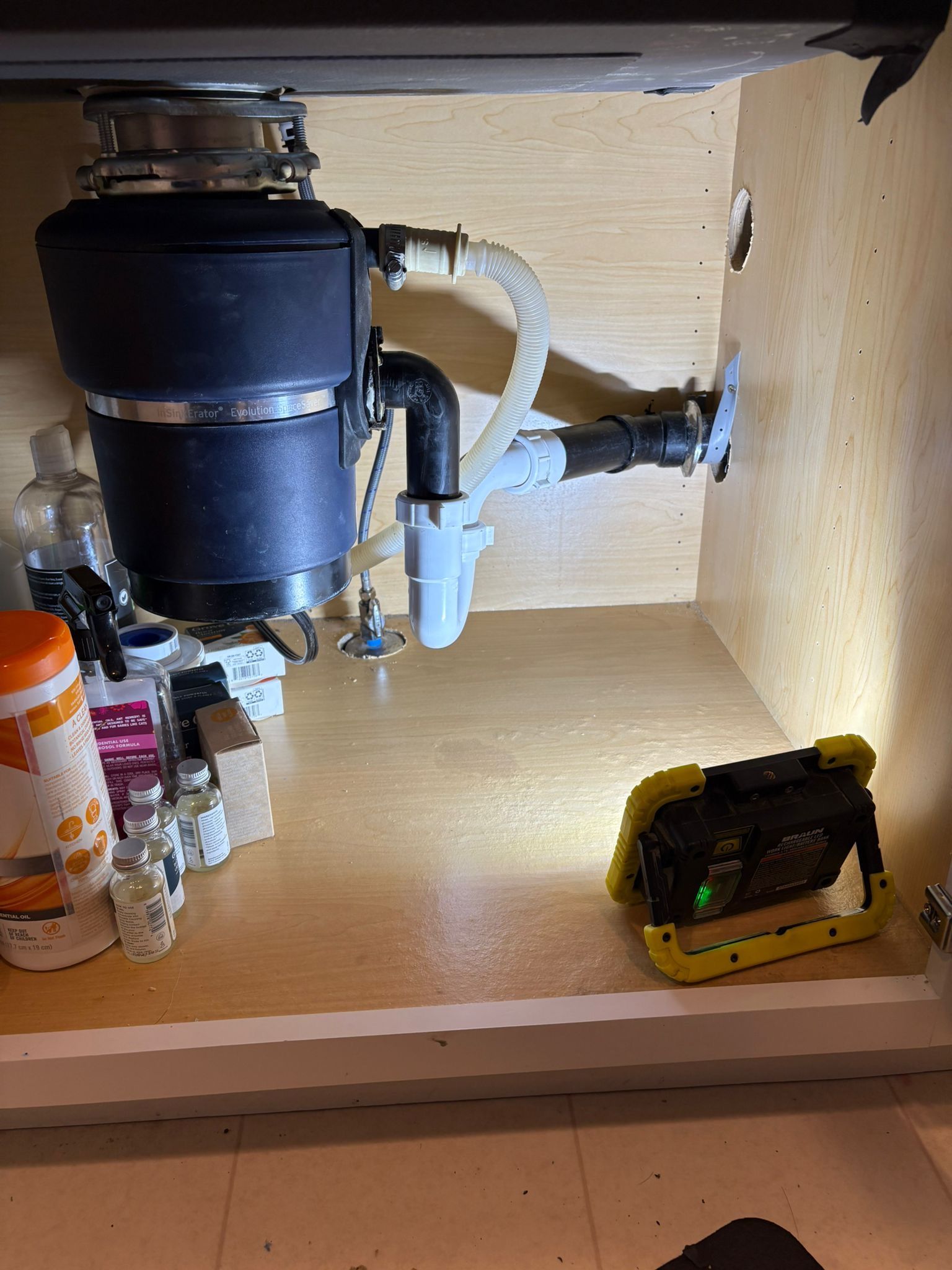 Garbage disposal and plumbing under a kitchen sink. A work light illuminates the space.
