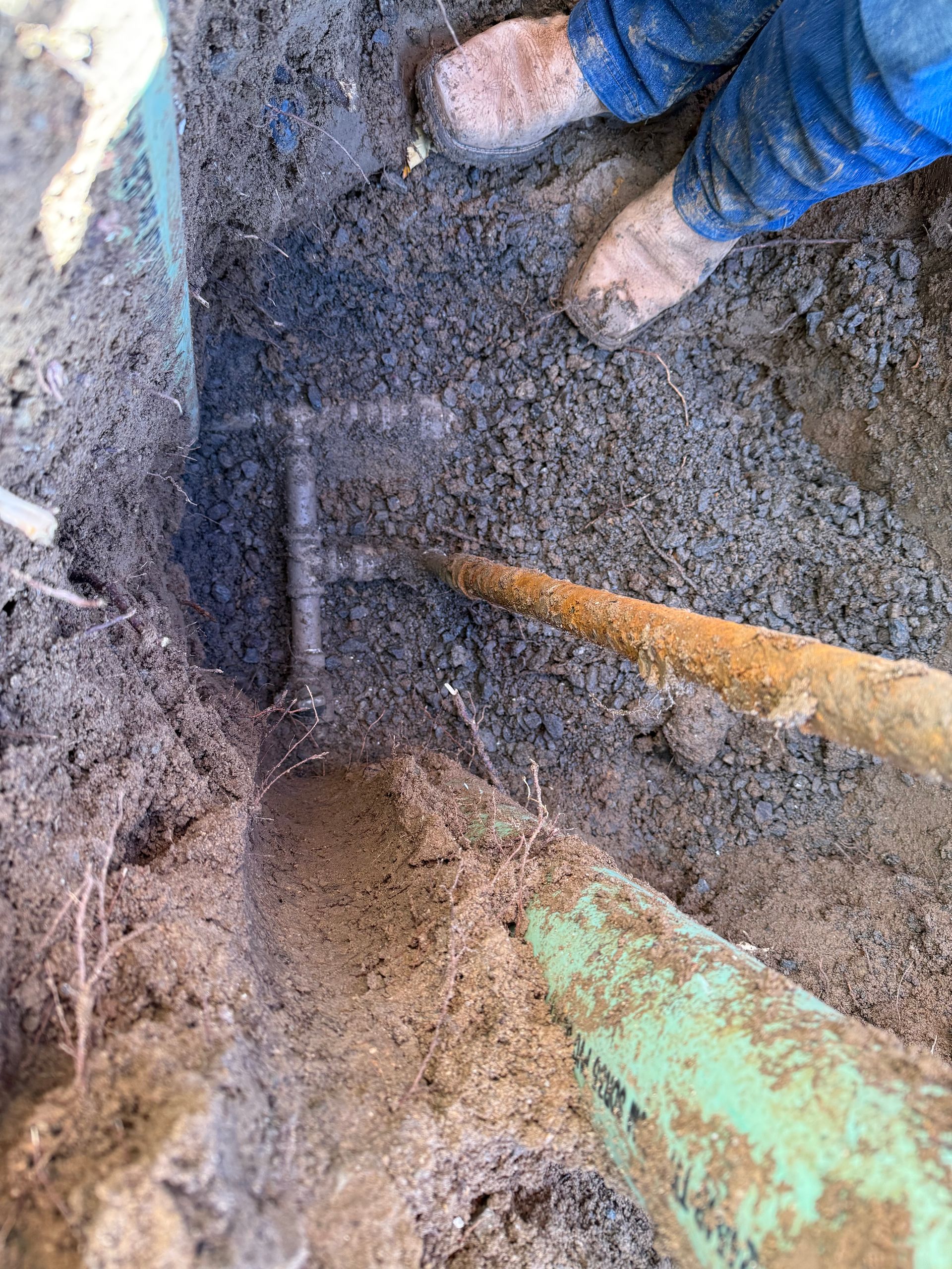 Person in blue pants and muddy boots repairs a buried pipe in a dirt trench, using tools.