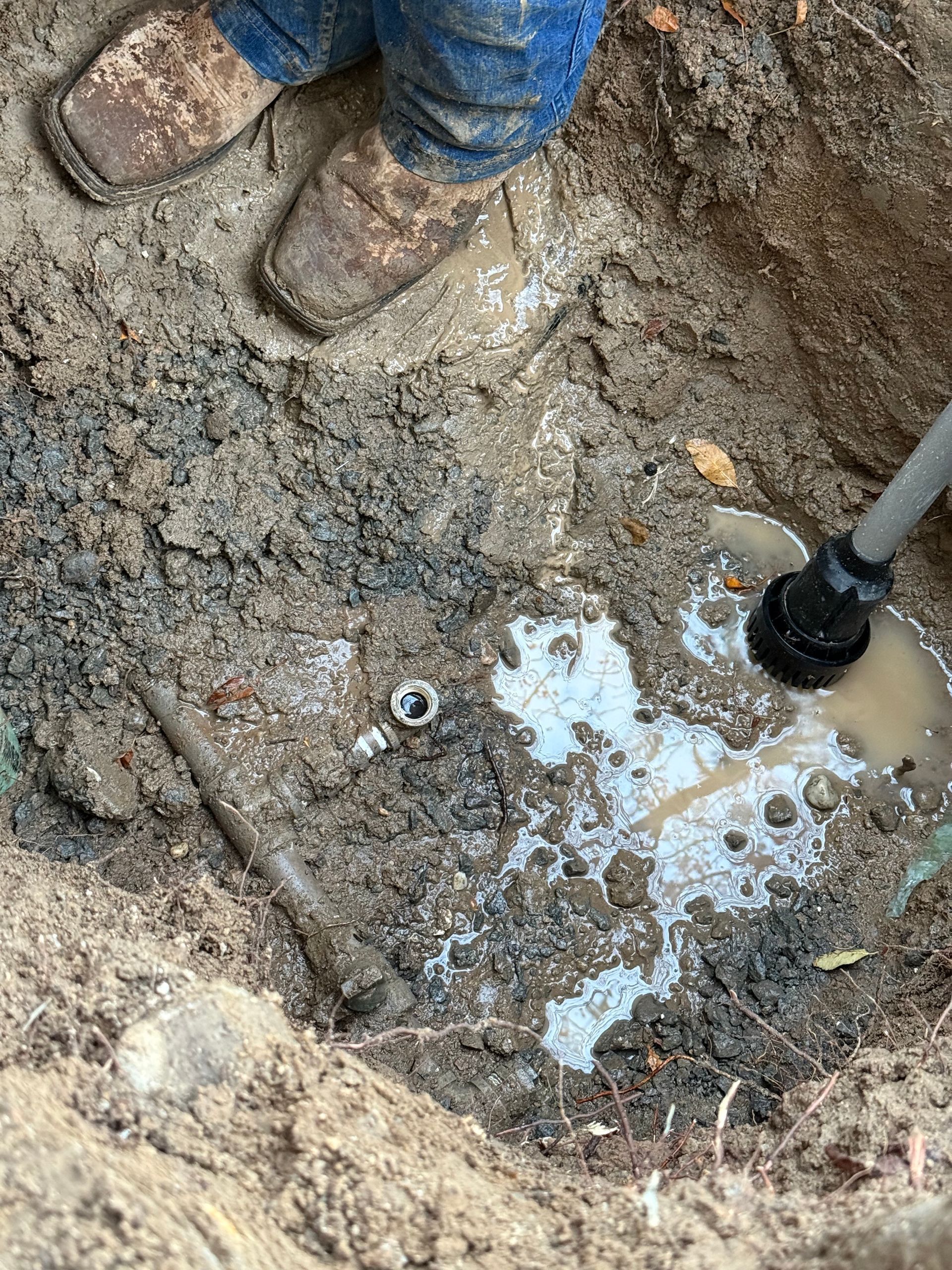 Person in muddy boots excavates a water pipe, surrounded by wet dirt and a pool of water.