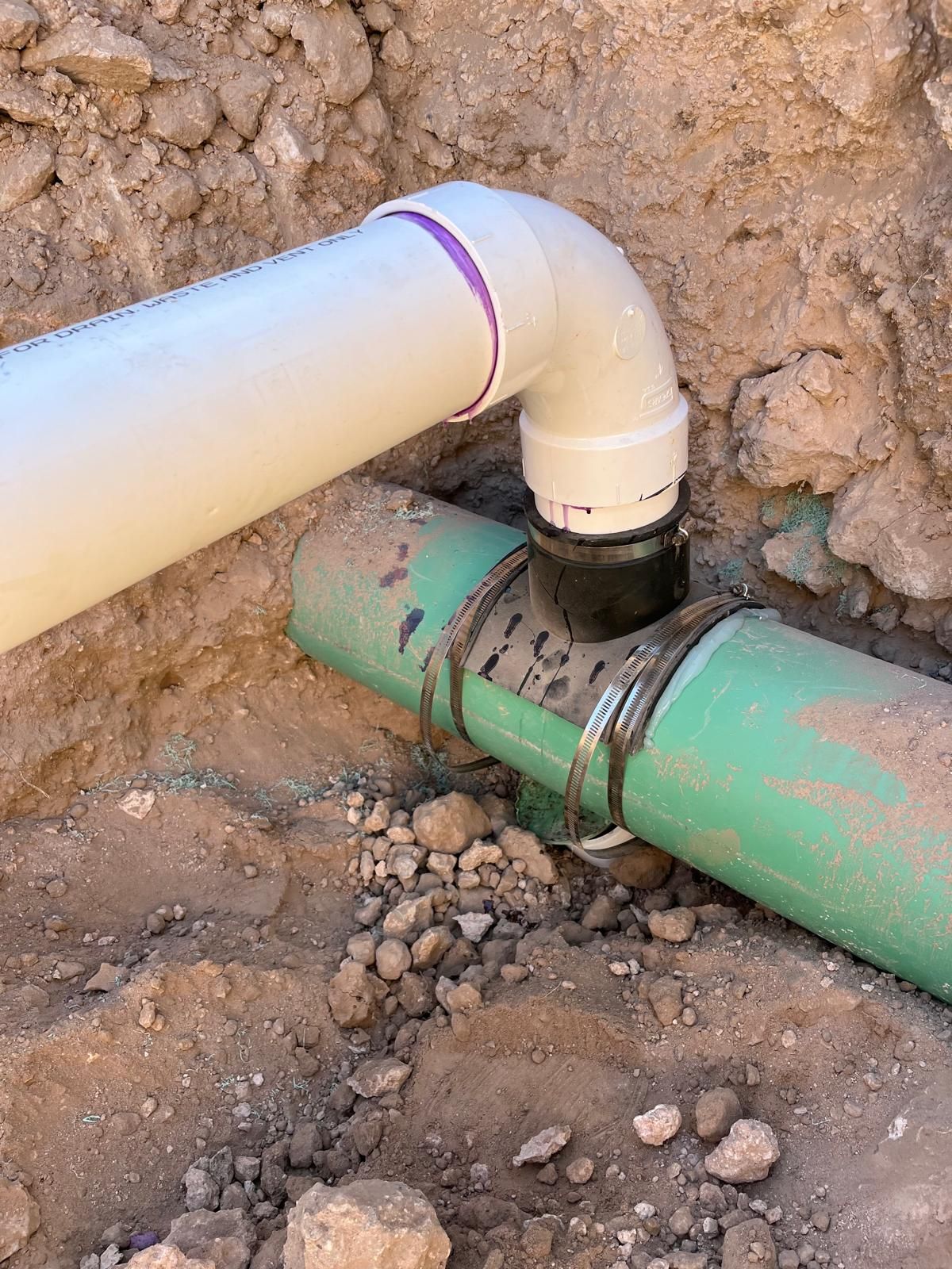 White PVC pipe connected to green pipe in a dirt trench, using a black fitting.