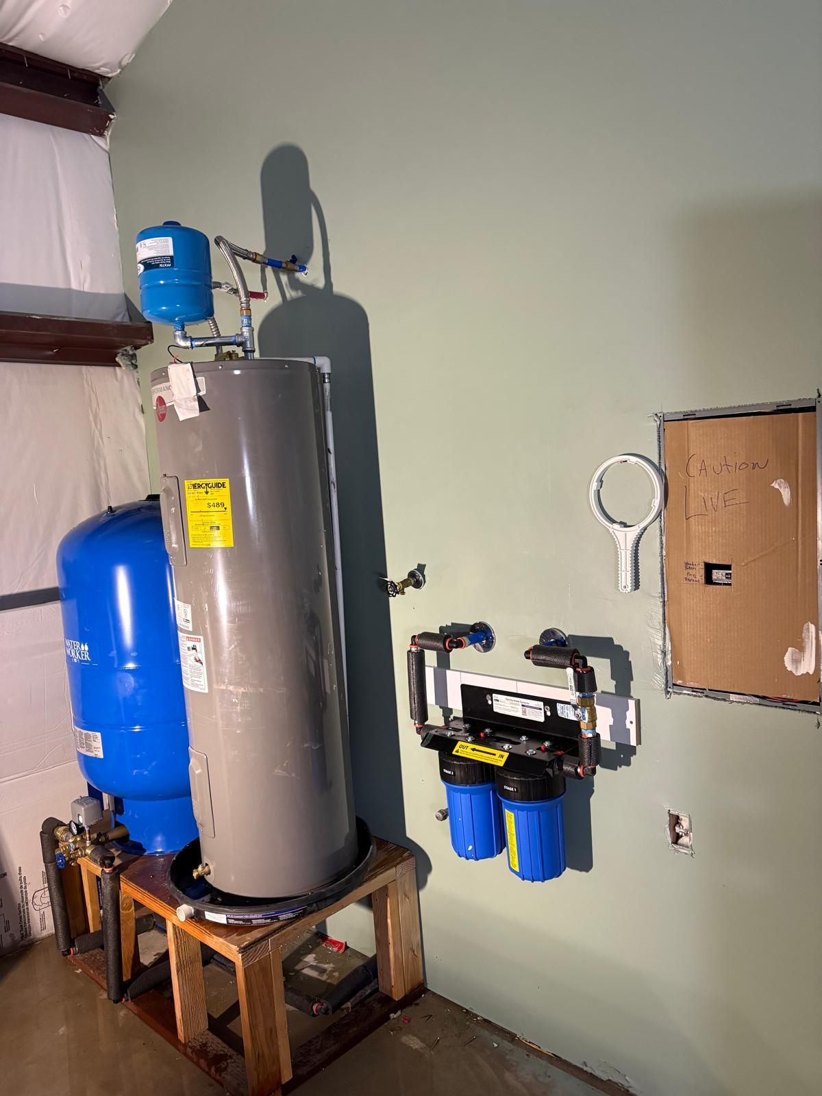 Water well equipment against a green wall: blue pressure tank, water heater, filter, and control panel.