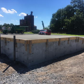 Concrete foundation pad at a construction site with trucks, silo, and trees under a sunny blue sky
