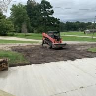 Orange skid steer leveling dirt on a roadside lot near a driveway and trees