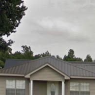 Single-story house with gray metal roof under a cloudy sky, framed by trees.