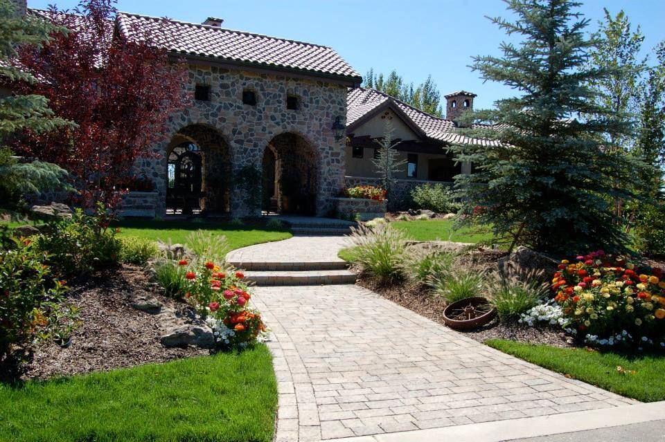 A stone path leads to the arched entryway of a rustic, multi-story stone house with a tiled roof and landscaped yard.