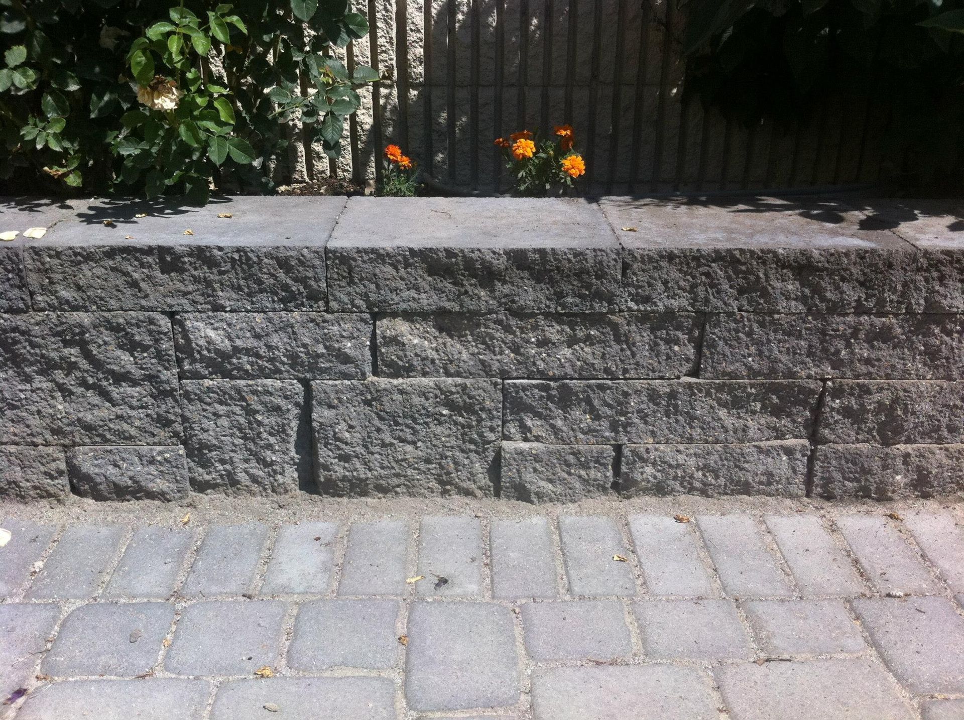 A low, gray concrete retaining wall made of stacked, textured blocks, set against a background of greenery and paving.