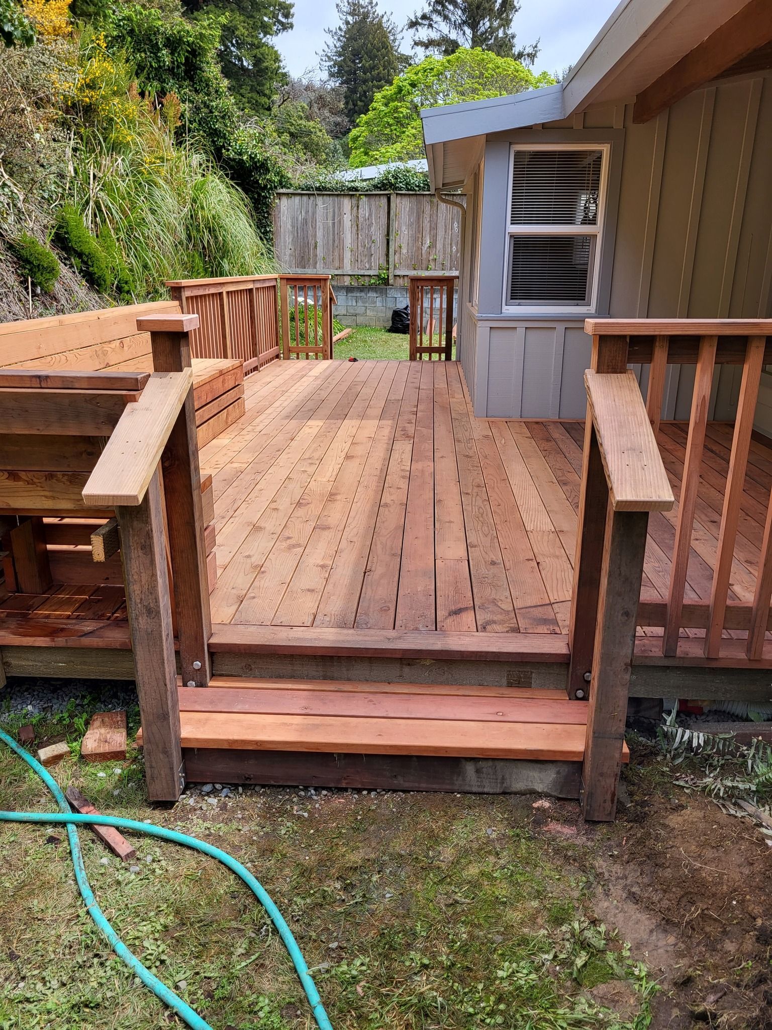 A wooden deck with stairs leading up to it and a hose in the grass.