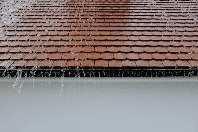 Rainwater flowing from a residential shingle roof during heavy rainfall with visible roof drainage and runoff.