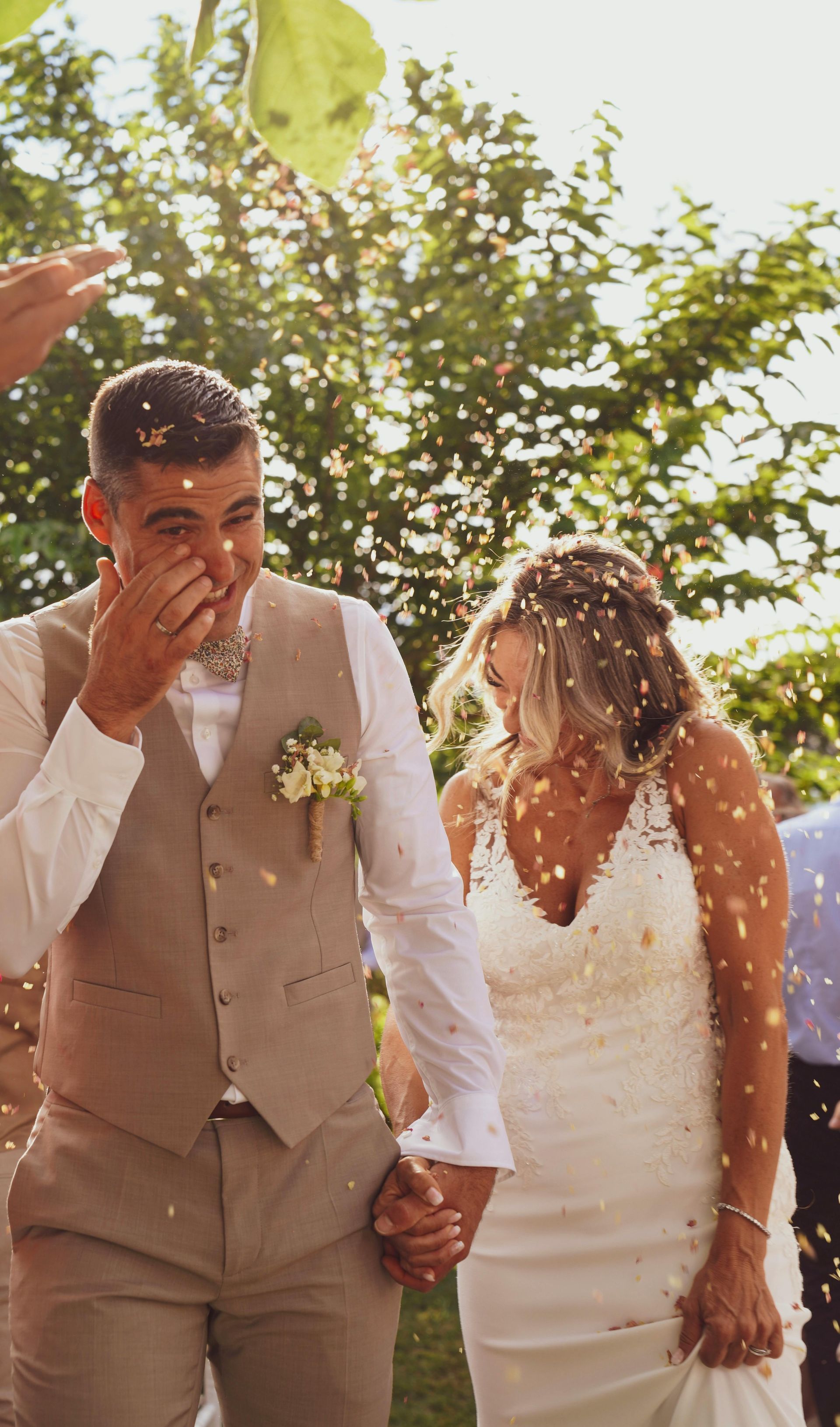 A bride and groom walk hand-in-hand outdoors through falling confetti, smiling with their eyes closed.