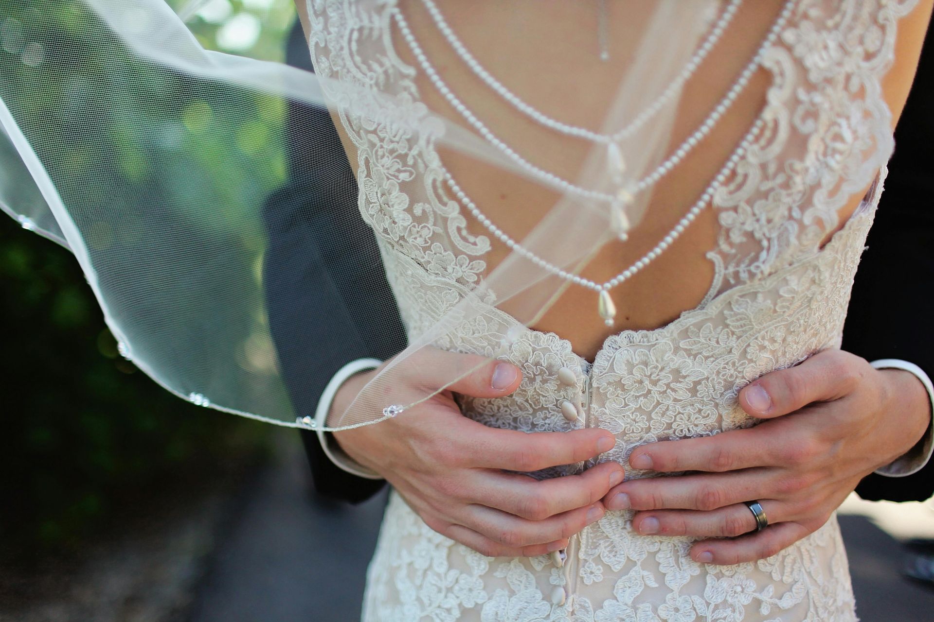 Close-up of a person embracing another wearing a lacy, pearl-embellished wedding dress with a sheer veil blowing outdoors.