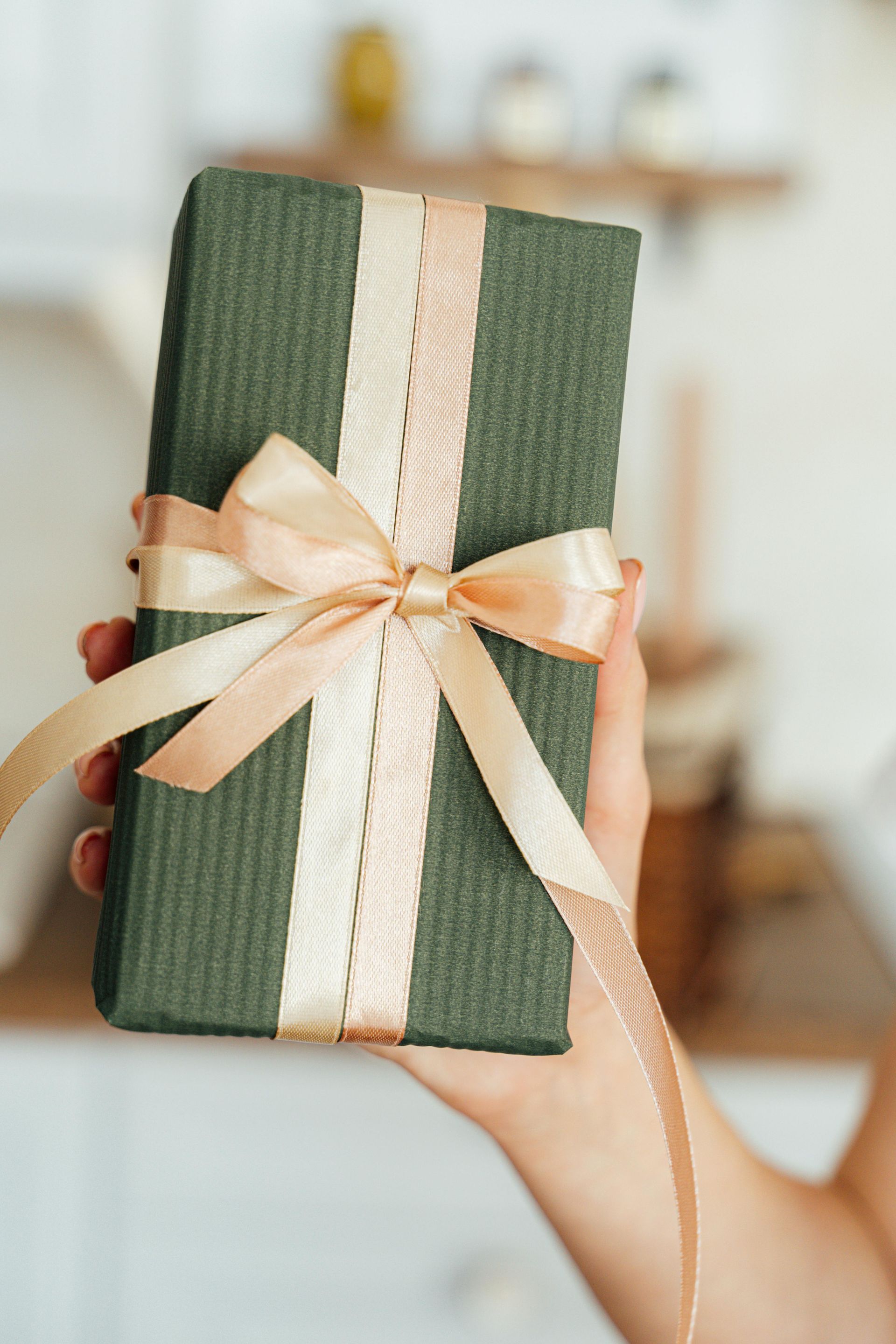 A hand holds a gift box wrapped in dark green, textured paper with a wide, light-colored ribbon tied in a bow.