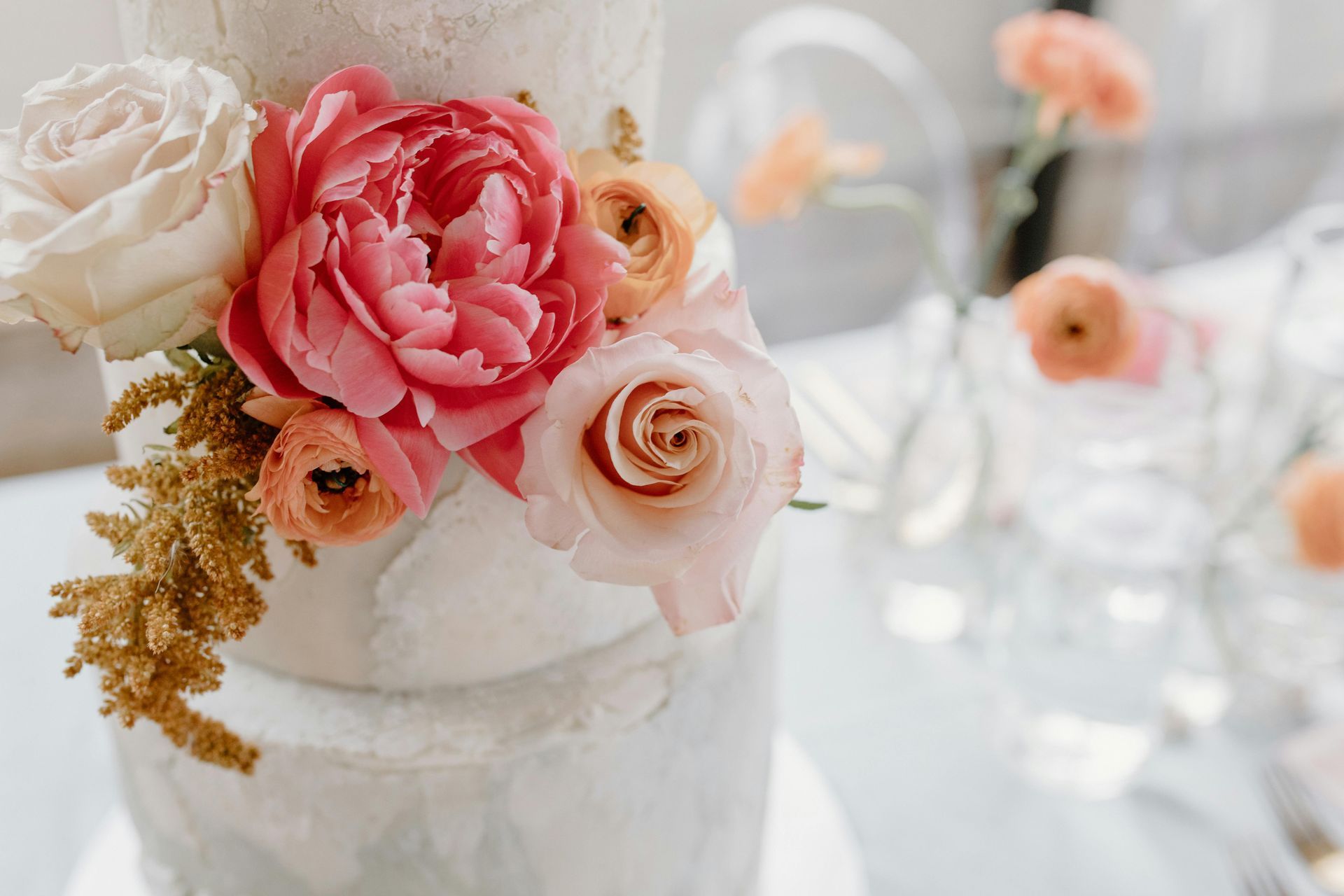 A multi-tiered white textured cake decorated with vibrant pink, peach, and white flowers and golden greenery.