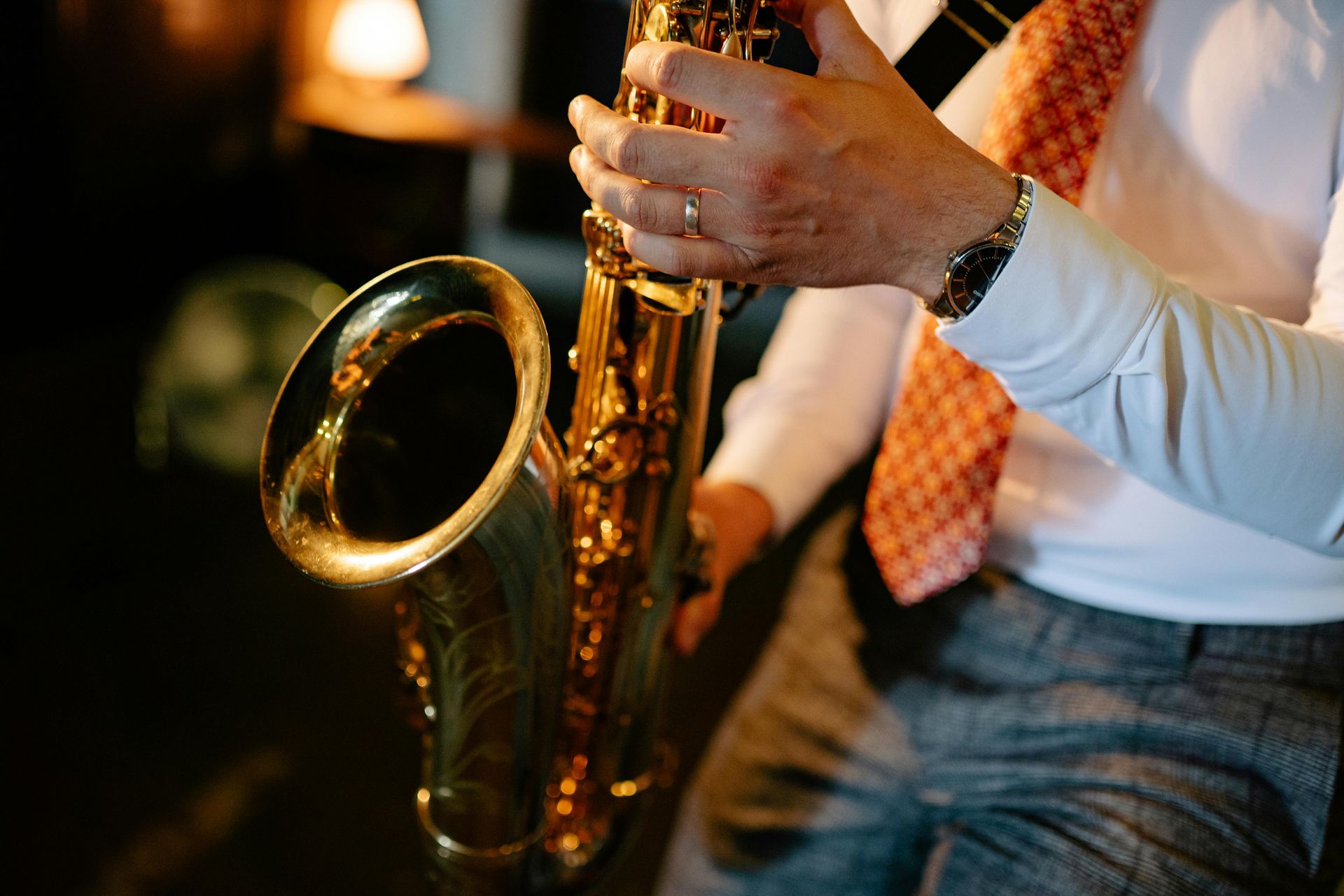 A person in a white dress shirt and patterned orange tie plays a gold saxophone, with their hand resting on the keys.